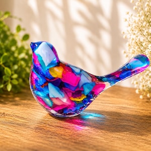 May include: A colorful glass bird figurine. The bird is made of fused glass in shades of blue, pink, yellow, and white. The figurine is sitting on a wooden surface.