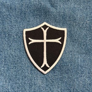 May include: A black and white shield-shaped patch with a cross design. The patch has a black background with a white cross and a white border. The patch is on a blue denim background.
