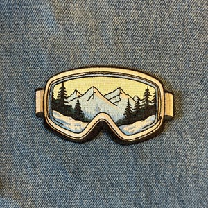 May include: Embroidered patch featuring a pair of ski goggles with a mountain and forest scene inside. The patch has a cream-colored border and is set against a blue denim background. The goggles' lenses depict a snowy mountain landscape.