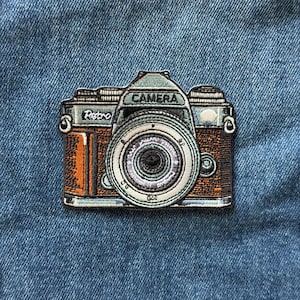 May include: An embroidered patch of a camera with a brown body and light blue accents. The word Retro is stitched in white on the left side, and the word CAMERA is stitched in black on the top center of the camera body. The lens is circular.