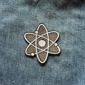 May include: An embroidered patch featuring a dark gray atom symbol with a white center circle and white orbital rings. The design includes three small white circles positioned along the orbital paths, all outlined in a thick black border.