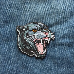 May include: An embroidered patch featuring the head of a snarling black panther with an open mouth, sharp white fangs, a pink tongue, and piercing orange eyes. The design includes detailed stitching in shades of black, grey, and light blue.