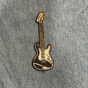 May include: Embroidered patch of an electric guitar. The patch features a black body with white accents, a brown neck, and a yellow headstock. The guitar patch is likely for clothing or accessories.