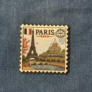 May include: A rectangular embroidered patch shaped like a postage stamp featuring the Eiffel Tower, the Sacré-Cœur Basilica, a bridge over the Seine, and pink cherry blossoms. The text reads PARIS FRANCE with a circular postmark stamp.