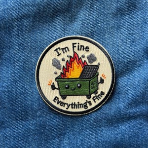 May include: A circular embroidered patch with a beige background and a black border. It features a green dumpster on fire with a smiling face, giving a thumbs up with both hands. The text reads I'm Fine at the top and Everything's Fine at the bottom.