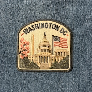 May include: A rectangular embroidered patch with rounded top corners featuring the text WASHINGTON DC in black. The design depicts the US Capitol building, the Washington Monument, a waving American flag, and pink cherry blossom branches.