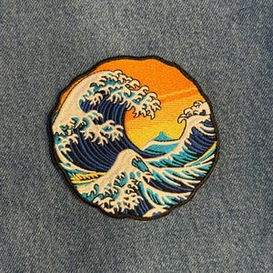 May include: Embroidered patch featuring a stylized wave design with a setting sun in shades of orange and yellow. The wave is depicted in shades of blue and white, with a black border. The patch is circular and set against a denim background.