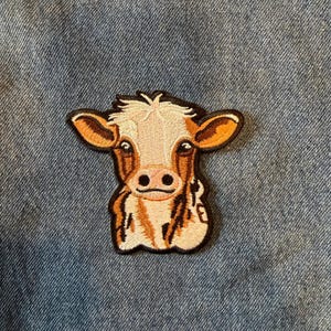 May include: Embroidered patch featuring a cartoon cow face. The cow has a white face with brown and tan markings, brown ears, and a black outline. The patch is likely for clothing or accessories.