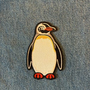 May include: Embroidered penguin patch with a white belly, black wings, and a gray head. The penguin has a yellow chest, red feet, and a red beak. The patch is outlined in black and is on a blue denim background.