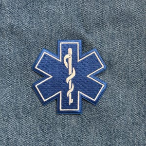 May include: A blue embroidered Star of Life patch with a white border and a white staff of Asclepius in the center. The patch is on a denim background. This patch is commonly used to identify emergency medical services.