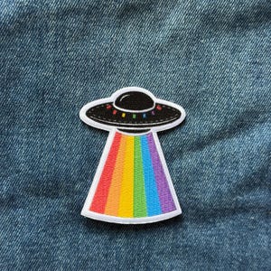May include: An embroidered patch featuring a black UFO saucer with small colorful dots along the rim, emitting a wide, vertical beam of light composed of vibrant rainbow-colored stripes, all outlined in a clean white border against a blue denim fabric.
