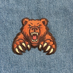 May include: Embroidered patch featuring a roaring bear with orange and brown fur, white teeth, and sharp claws. The patch is set against a blue denim background, showcasing the bear's aggressive expression.