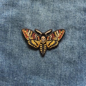 May include: An embroidered patch featuring a death's-head hawkmoth with a skull pattern on its thorax. The moth has wings in shades of yellow, orange, and black, with a black body and antennae, displayed against a blue denim fabric background.