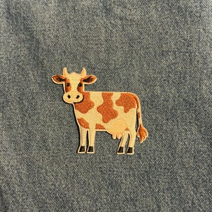 May include: Embroidered patch featuring a cartoon cow with brown and white spots. The cow has a cream-colored face and body, with black hooves and horns. The patch is designed to be ironed or sewn onto fabric.