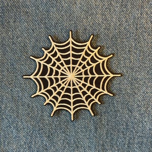 May include: A black and white spider web patch. The web design is white with a black background and a black border. The patch is placed on a blue denim background.