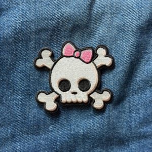 May include: An embroidered patch featuring a white skull with large black eye sockets and a small pink bow on top, positioned over two crossed light gray bones. The design is outlined in black thread and is attached to blue denim fabric.
