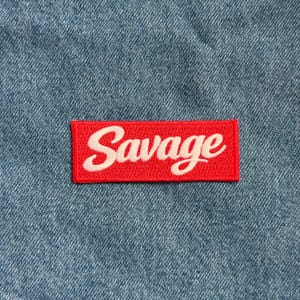 May include: A red rectangular patch with the word "Savage" embroidered in white script. The patch is set against a blue denim background, creating a contrast in color and texture.