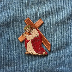 May include: An embroidered patch featuring a figure with long brown hair wearing a green crown and a red and white robe, kneeling while carrying a large brown wooden cross. The patch is stitched with detailed thread work and textured borders.