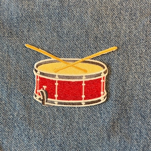 May include: A red drum patch with a yellow drumhead and two crossed drumsticks. The patch has a black and white border and is set against a blue denim background.