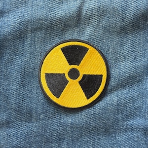 May include: A circular embroidered patch featuring a yellow background with a black radiation hazard symbol in the center. The design consists of three black blades radiating from a central black circle, all enclosed within a thick black border.