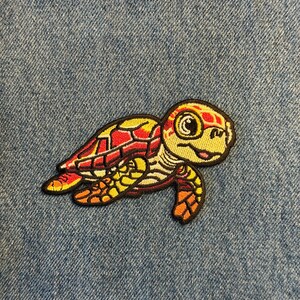 May include: Embroidered patch of a sea turtle in shades of red, yellow, and orange with a black outline. The turtle is smiling and has detailed stitching. The patch is on a blue denim background.
