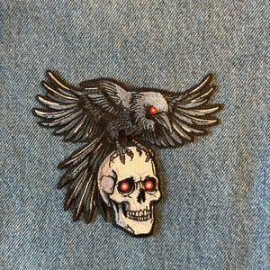 May include: Embroidered patch featuring a black raven with outstretched wings perched atop a white skull. The raven and skull have red eyes. The patch is on a blue denim background.