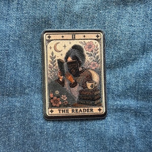 May include: A rectangular embroidered patch featuring a tarot card design titled THE READER. The illustration depicts a person reading a book, surrounded by flowers, a crescent moon, and a stack of books with a teacup, all in muted earth tones.