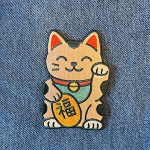 May include: Embroidered patch featuring a tan Maneki-neko cat with a raised paw, a green collar, and a gold coin. The cat has black spots and red accents on its ears. The patch is on a blue denim background.