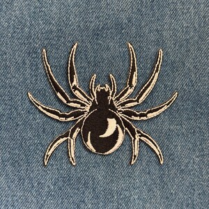 May include: Black spider patch with white embroidered details. The spider has a rounded body and eight legs. The patch is on a blue denim background.