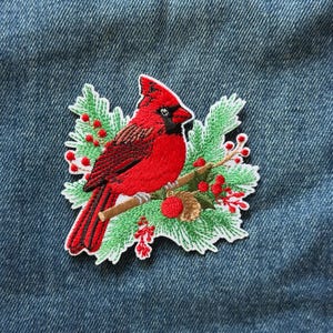 May include: An embroidered patch featuring a bright red cardinal bird perched on a brown branch. The bird is surrounded by green pine needles, small red berries, and a single red ornament. The patch is placed against a blue denim fabric background.