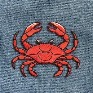 May include: A red crab patch with black embroidered details, including claws and legs, is centered on a blue denim background. The crab's claws are raised, and the patch is likely for clothing or accessories.