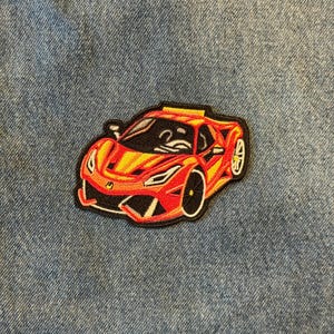 May include: Embroidered patch featuring a red and yellow sports car with black wheels and a black outline. The patch is on a blue denim background. The car design is detailed and stylized.
