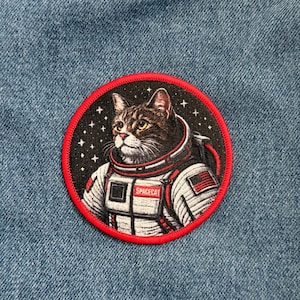 May include: Embroidered patch featuring a tabby cat in a spacesuit, set against a starry black background within a red circular border. The patch includes the text "SPACECAT" and an American flag.