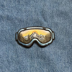 May include: Embroidered patch depicting a pair of ski goggles with a mountain scene. The goggles are outlined in blue and black, with a sunset over snow-capped mountains and trees visible through the lenses. The patch is on a blue denim background.
