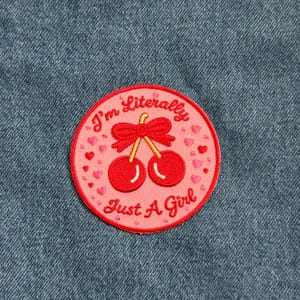 May include: A round, pink embroidered patch with a red border. The patch features a pair of red cherries with a bow, surrounded by hearts. Text on the patch reads "I'm Literally Just A Girl."