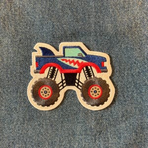 May include: Embroidered patch featuring a blue monster truck with a shark fin, red accents, and large black tires. The truck has a green windshield and a white border. This patch is perfect for decorating clothing or accessories.