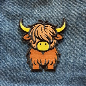 May include: An embroidered patch featuring a cartoon Highland cow with shaggy brown fur, a bright yellow muzzle, and curved yellow horns. The design is outlined in black thread and is positioned against a textured blue denim fabric background.