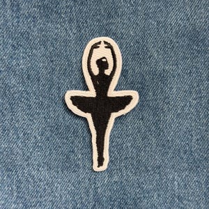 May include: Embroidered patch featuring a black ballerina silhouette with a white outline. The ballerina is in a graceful pose with arms raised above her head. The patch is on a blue denim background.
