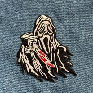 May include: Embroidered patch featuring the Ghostface character from the Scream horror film series. The patch is black and white with a red-stained knife. The patch is on a blue denim background.