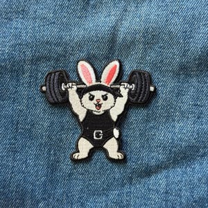 May include: An embroidered patch featuring a white rabbit with pink-lined ears wearing a black headband and a black weightlifting singlet. The rabbit is lifting a black barbell with large weights above its head, set against a blue denim fabric.