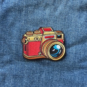 May include: A rectangular embroidered patch featuring a vintage-style camera with a bright red body, gold-toned accents, and a prominent lens. The camera design includes a black lens center with blue highlights and a small red rectangular detail.
