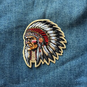 May include: An embroidered patch featuring a profile view of a person wearing a traditional feathered headdress. The design includes red, white, black, and tan thread colors, with detailed stitching depicting facial features and feathers.