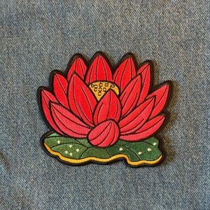 May include: Embroidered patch featuring a vibrant red lotus flower with yellow center details and green leaves. The patch has a black border and is set against a blue denim background. The lotus flower is a symbol of purity and enlightenment.