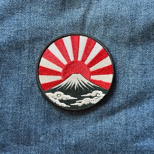 May include: A circular embroidered patch featuring a stylized mountain peak in dark green with white snow at the summit. Above the mountain, a red sun radiates white beams against a circular border. White clouds are stitched at the base of the mountain.