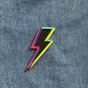 May include: A black lightning bolt patch with a colorful outline in red, yellow, blue, and green. The patch is embroidered and set against a blue denim background.