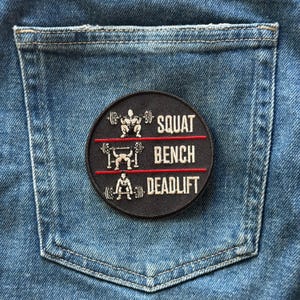 May include: A circular black embroidered patch featuring three white graphic illustrations of a person lifting a barbell. The text reads SQUAT, BENCH, and DEADLIFT, separated by two horizontal red lines. The patch is placed on blue denim fabric.