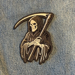 May include: Embroidered patch featuring the Grim Reaper in a black hooded cloak, holding a scythe. The patch has a skull face and skeleton hands. The design is in shades of black, white, and gray, with a stitched border. The patch is on a blue denim background.