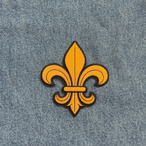 May include: Embroidered patch featuring a gold fleur-de-lis design with black outlining. The patch is set against a blue denim background. The fleur-de-lis is a stylized lily, a symbol often associated with royalty and heraldry.
