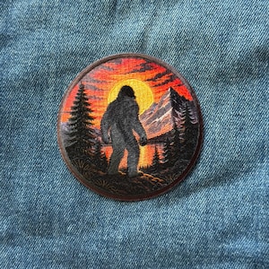 May include: A circular embroidered patch featuring a dark silhouette of a Bigfoot creature standing in a forest landscape. The background displays a vibrant orange and red sunset, a bright yellow sun, tall pine trees, and jagged mountain peaks.