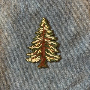 May include: Embroidered patch featuring a detailed evergreen tree design. The patch has a brown trunk, green needles, and white accents, set against a blue denim background. Ideal for adding a nature-inspired touch to clothing or accessories.